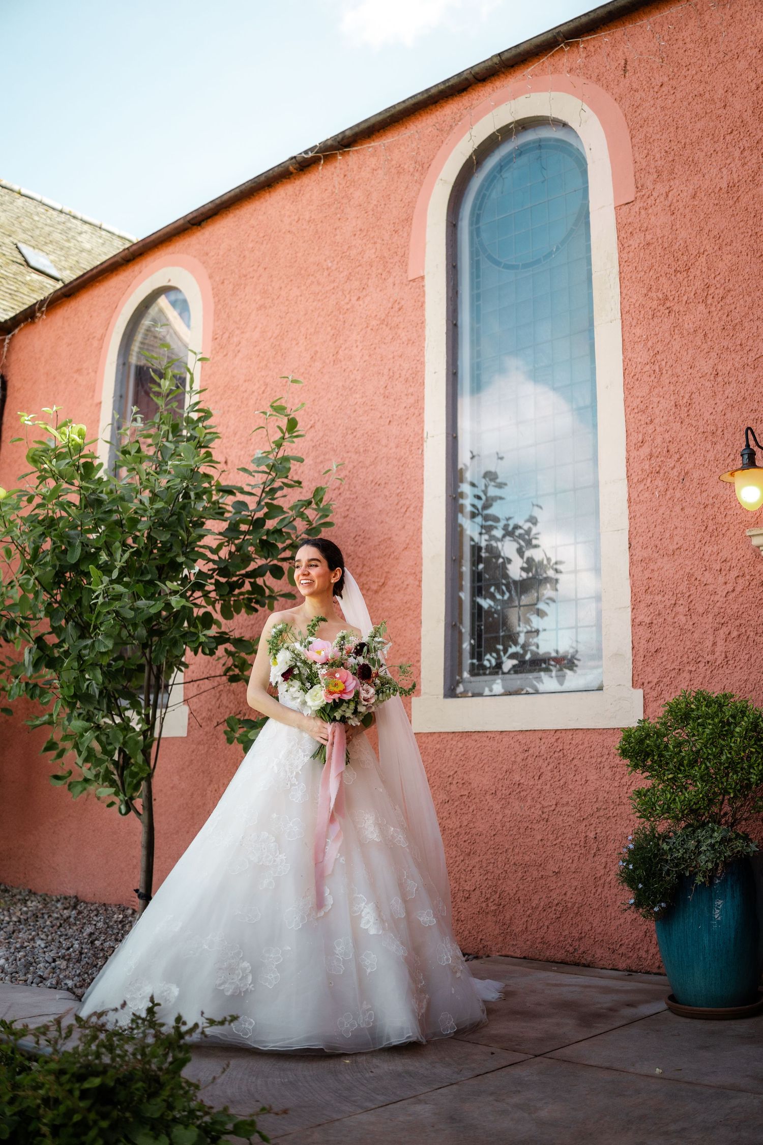 A bride in a flowing white gown poses against a coral-colored wall with arched windows and potted plants.