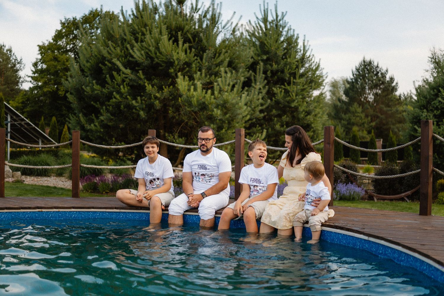 People sitting along the edge of a swimming pool with pine trees in the background.