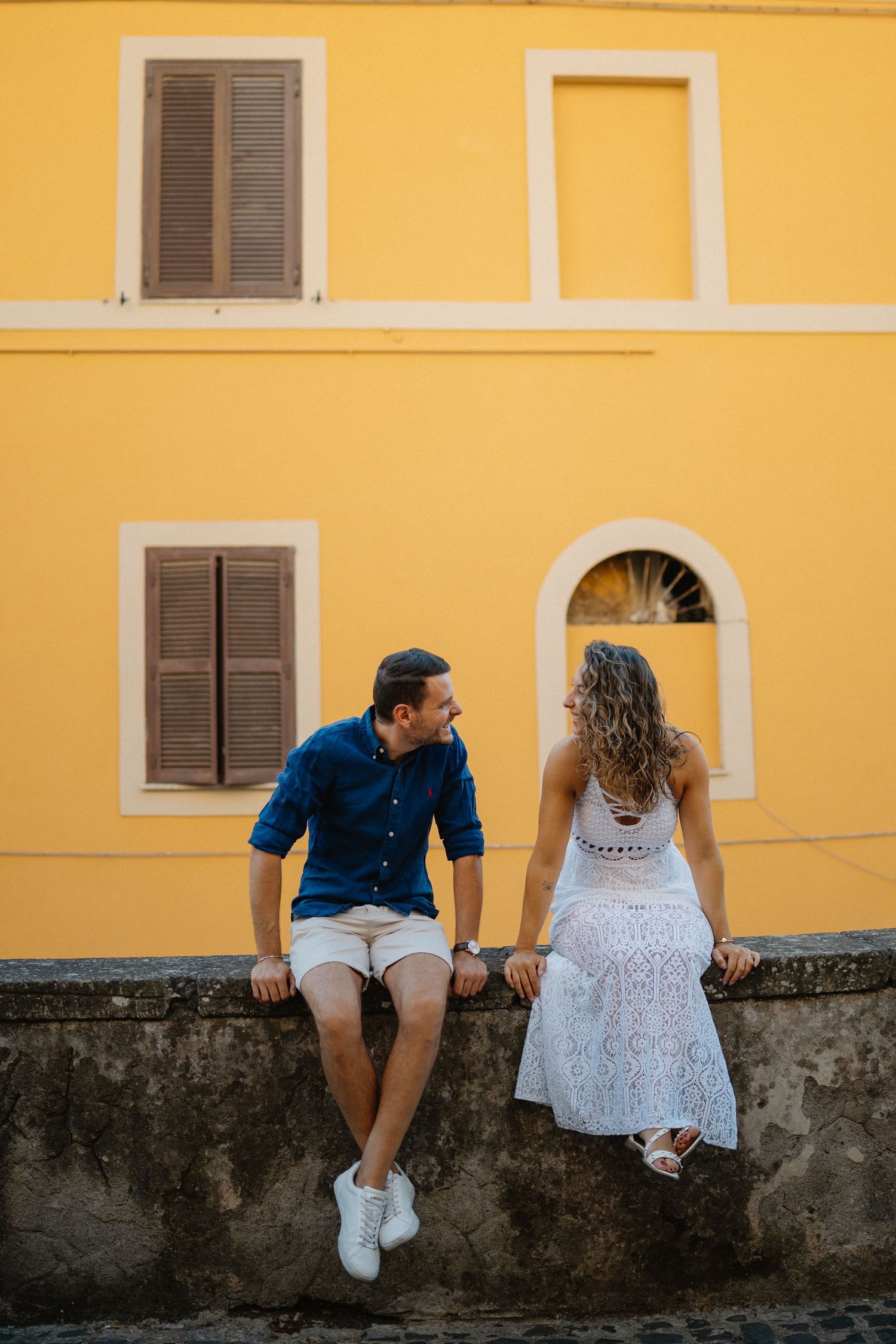 Couple sits casually on stone wall against bright yellow building with shuttered windows.