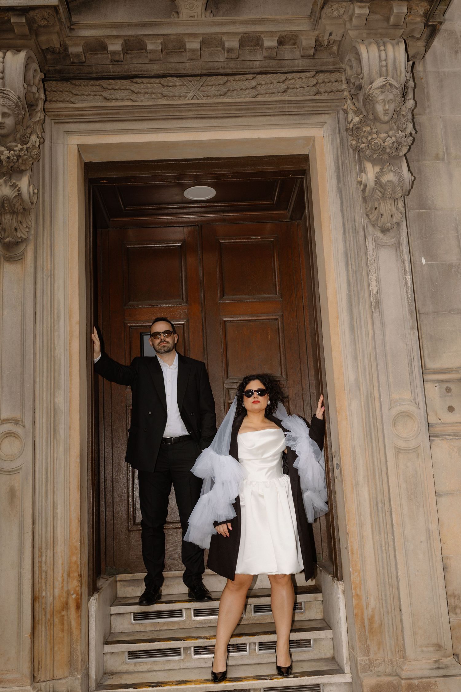 Stylish bride in short white dress and groom in black suit posing at ornate stone building entrance.