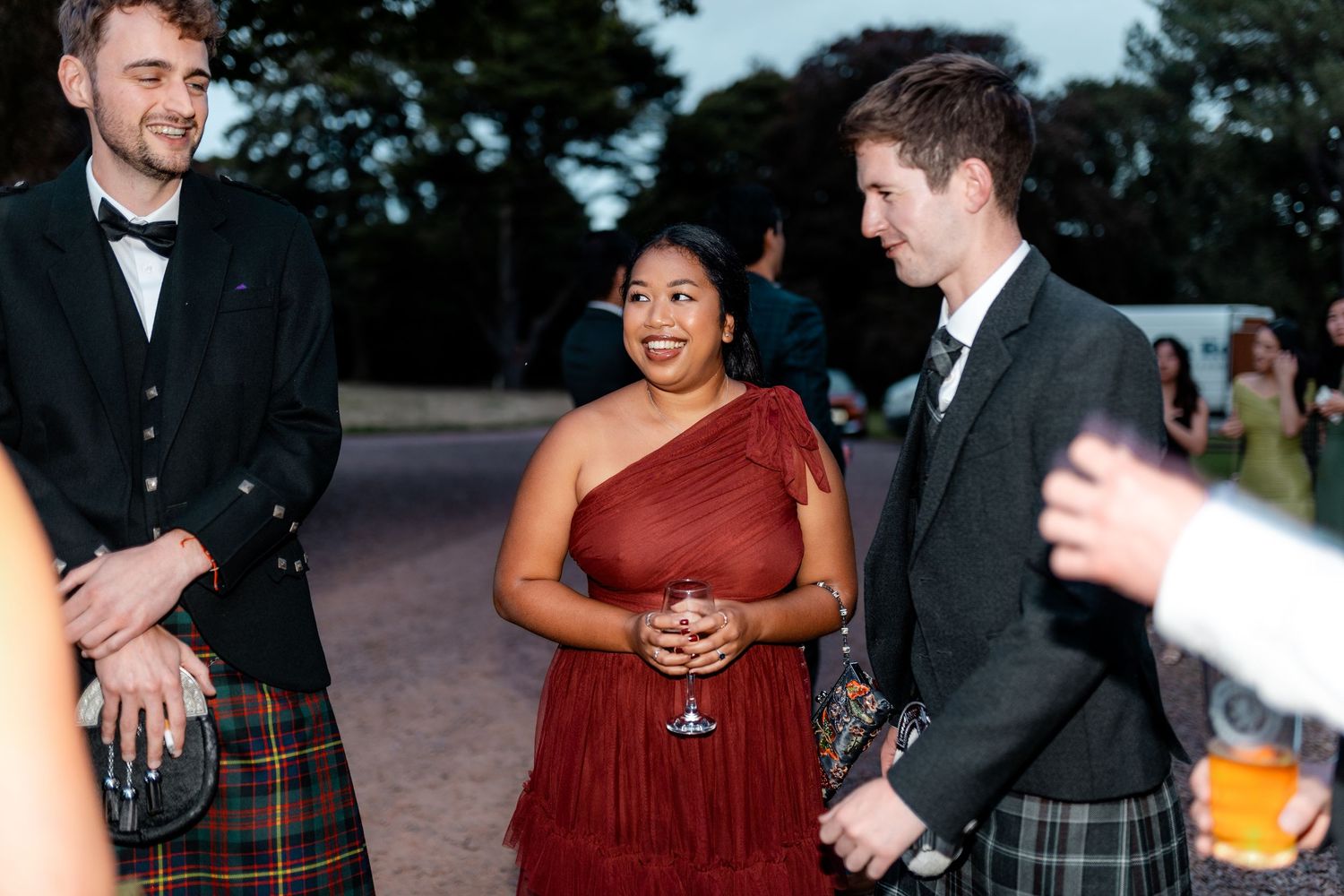 Two men in kilts and woman in red dress socializing outdoors at evening wedding reception.