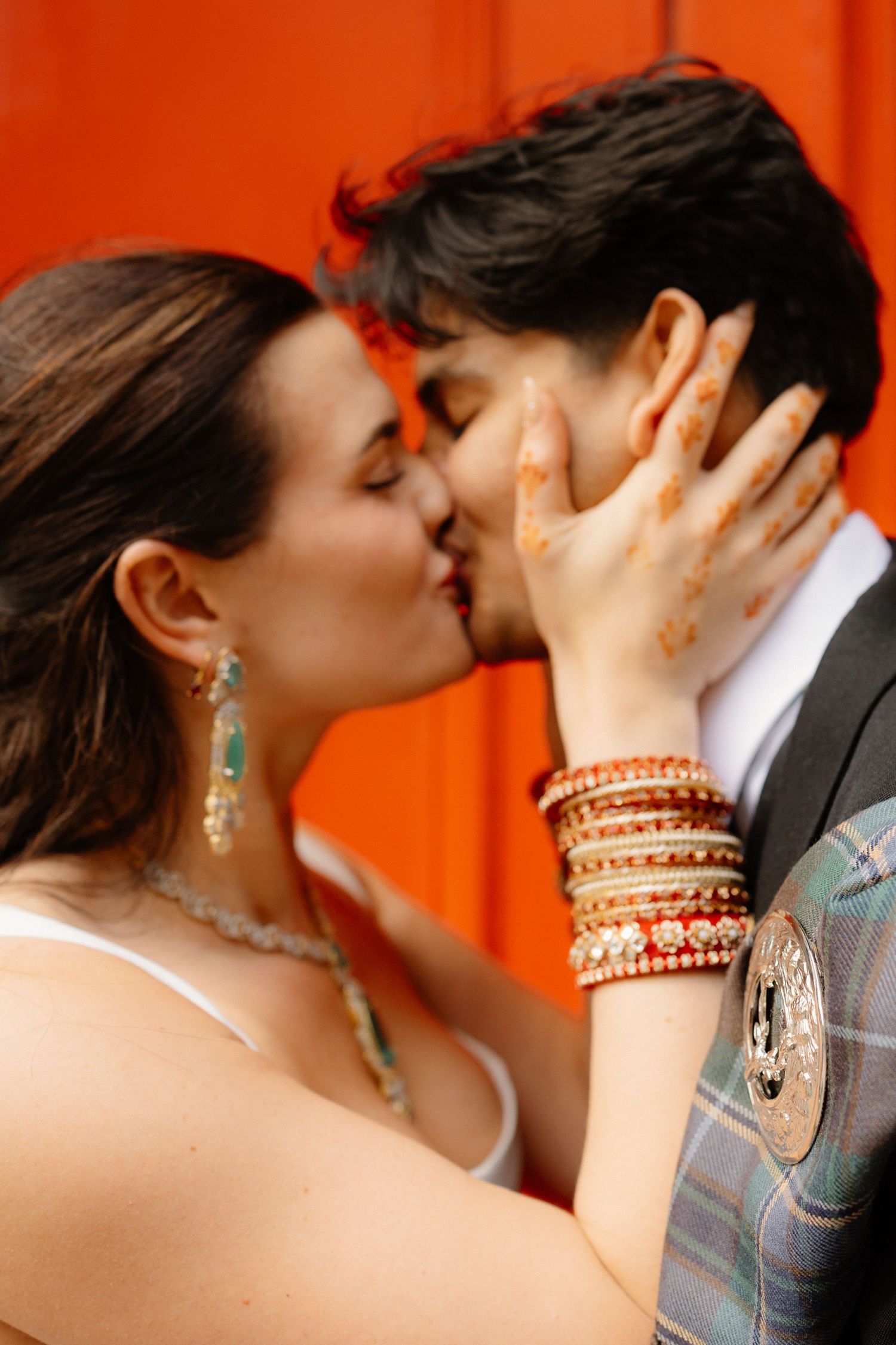 Couple sharing a romantic kiss with henna-adorned hands and colorful bangles against a vibrant orange backdrop.