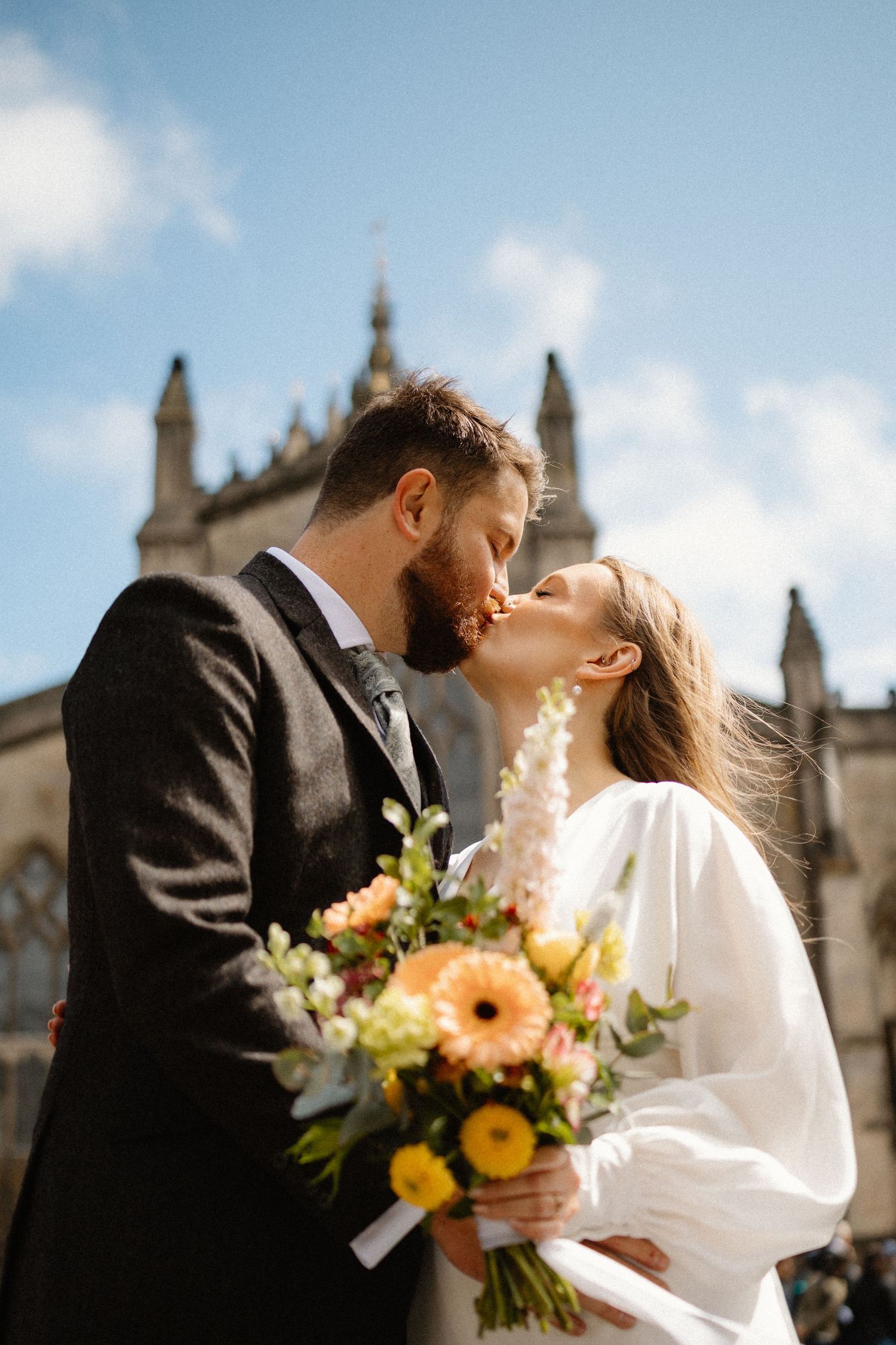A couple shares a romantic kiss outside a historic stone church, bride holding a vibrant bouquet of orange and yellow flowers.