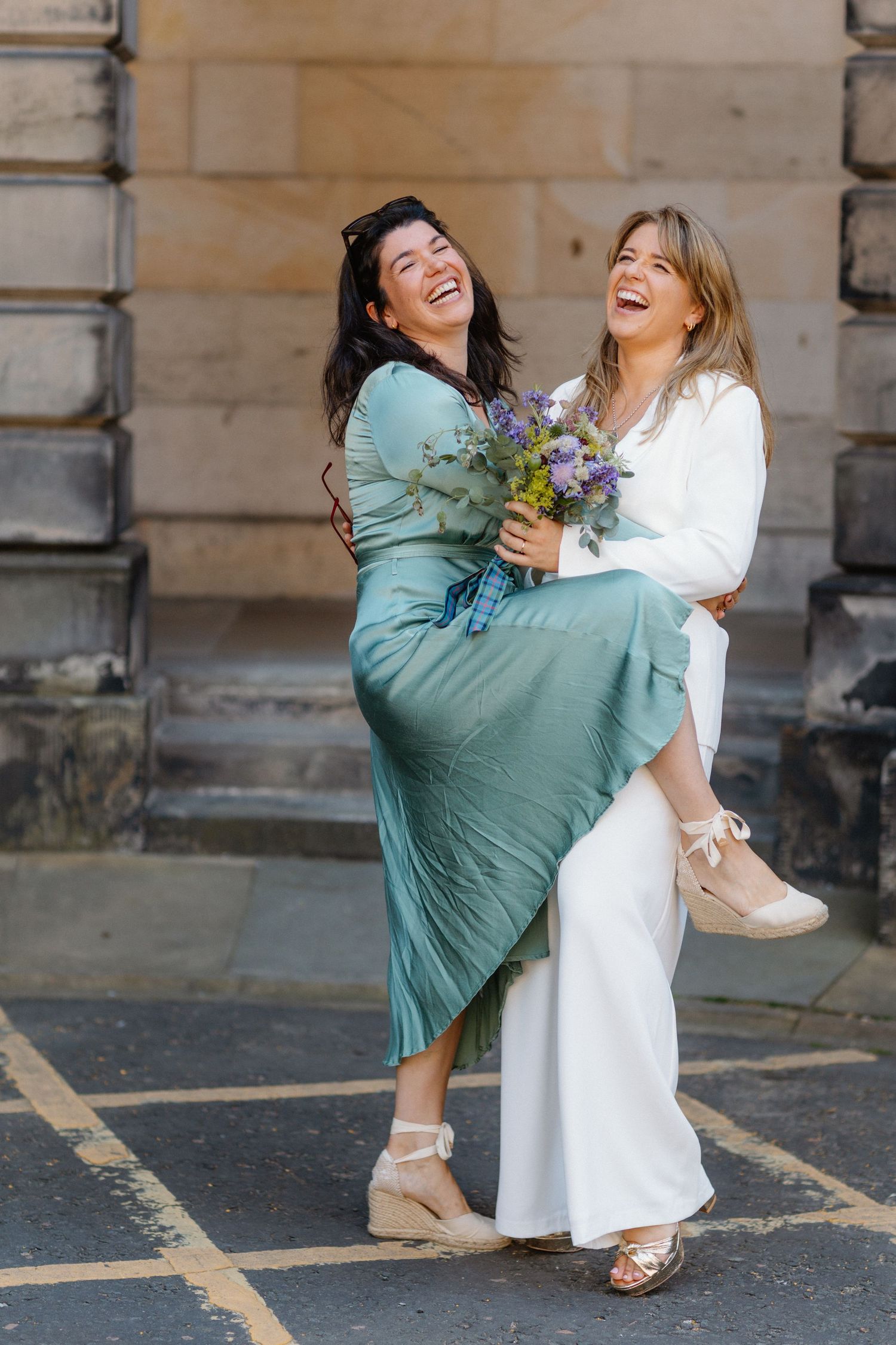 Two women laughing joyfully outdoors, one in white jumpsuit lifting bride in green dress with bouquet.