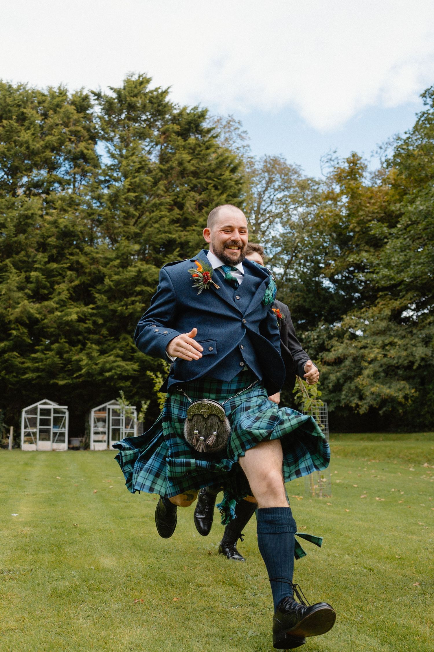 Joyful groom in navy jacket and blue tartan kilt running across green lawn at wedding.