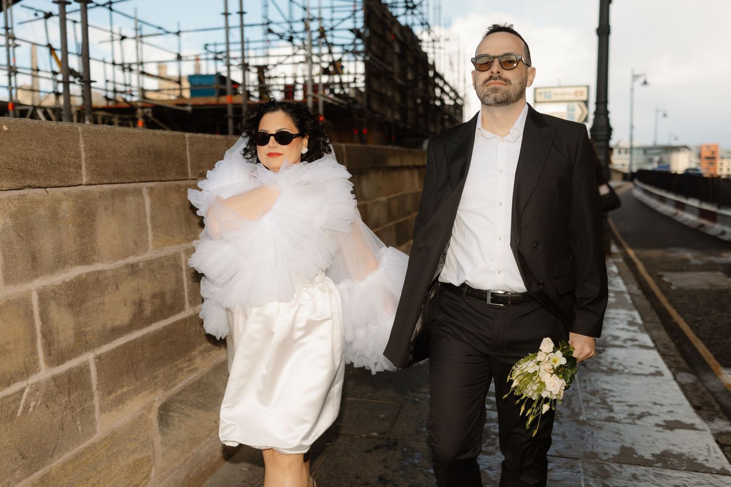 A bride in a white mini dress and groom in black suit hold hands on a cobblestone path near an industrial waterfront.