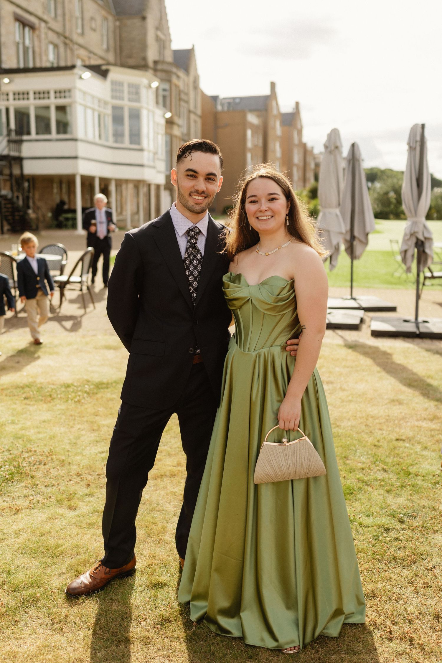 Couple posing outdoors at a formal event, man in dark suit, woman in elegant green strapless gown holding a cream clutch.