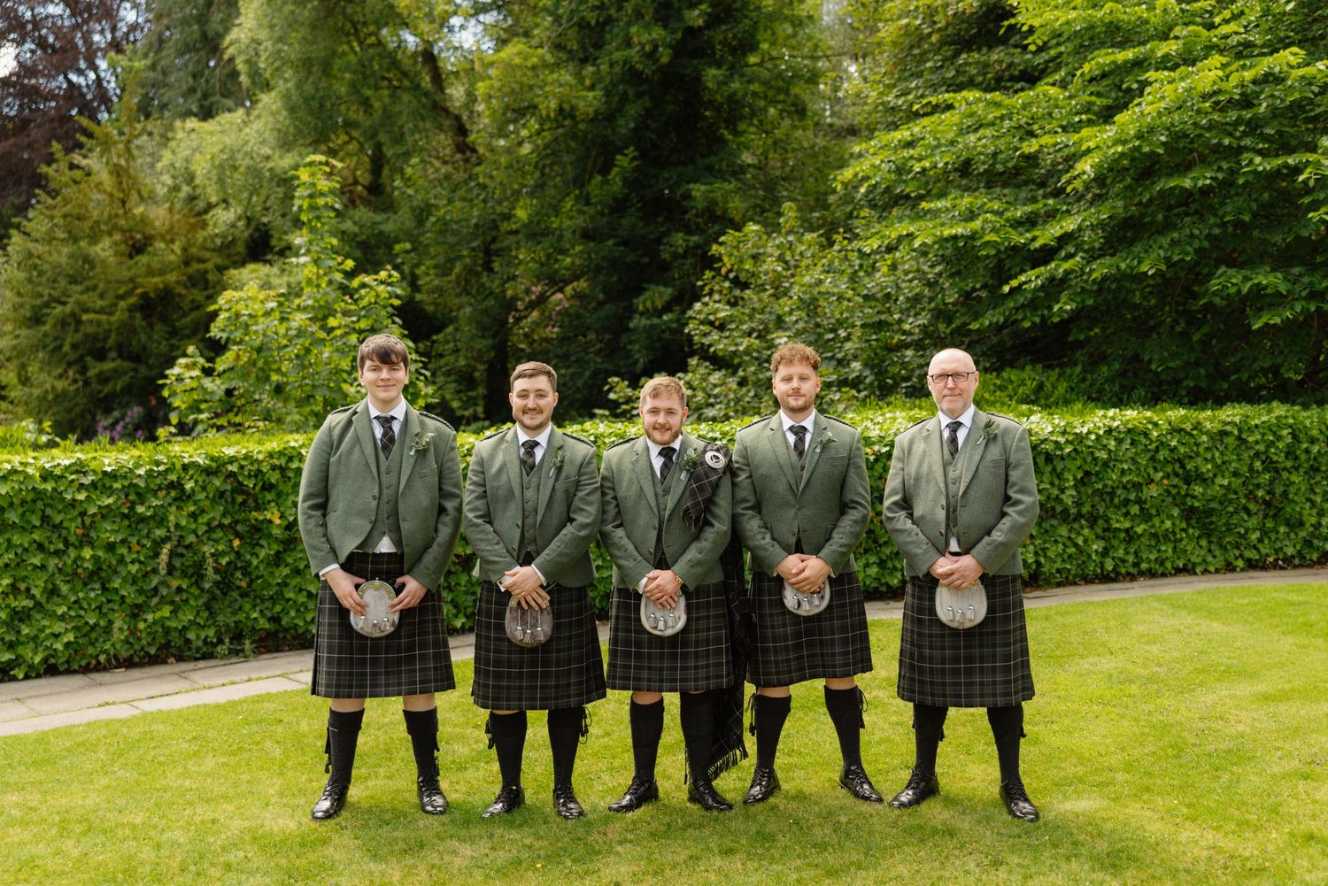 Five groomsmen in matching green tartan kilts posing outdoors in a garden at a Scottish wedding.