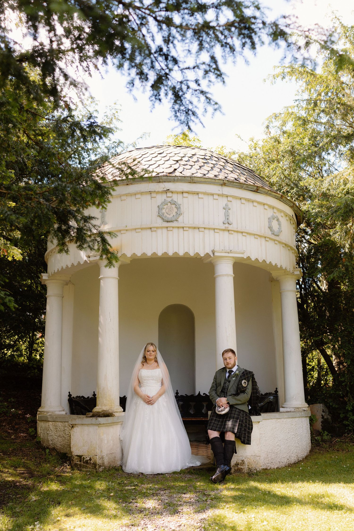 Bride in white gown and groom in Scottish kilt stand before a white classical gazebo surrounded by lush greenery.