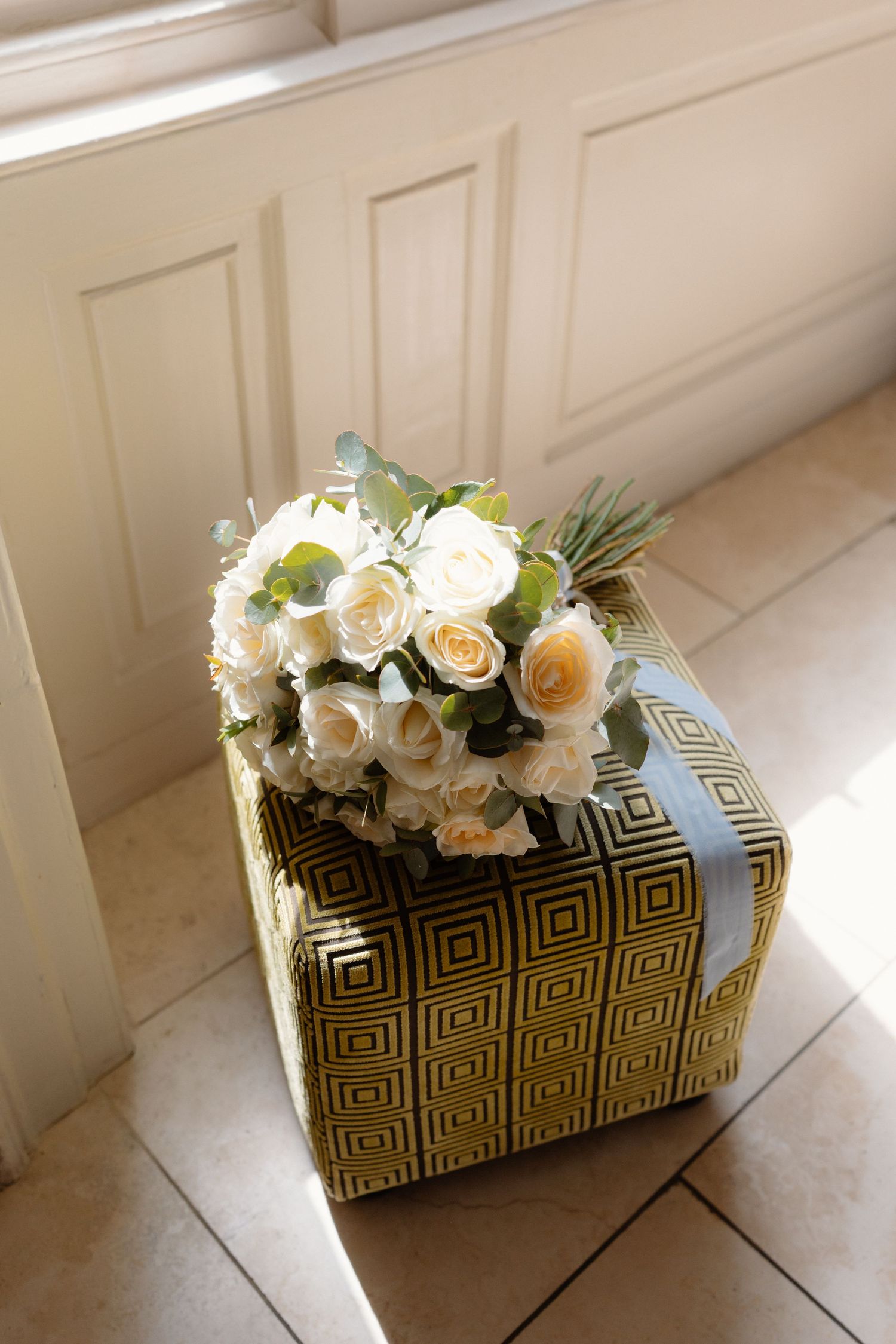 White and cream rose bouquet resting on a gold geometric patterned gift box with blue ribbon on tile floor.