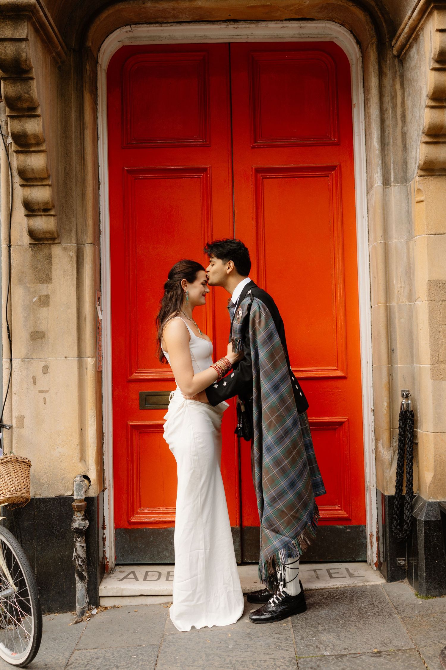 Bride in white dress and groom in Scottish tartan kilt sharing a romantic kiss before a vibrant red door.