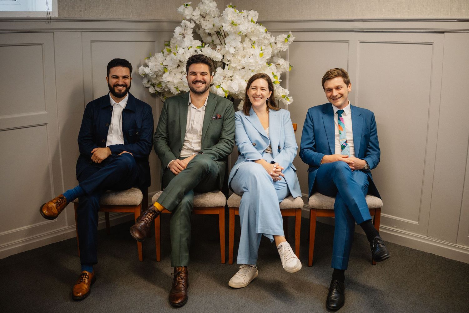 Group of people sitting on wooden benches in a casual indoor setting with white walls and natural lighting.