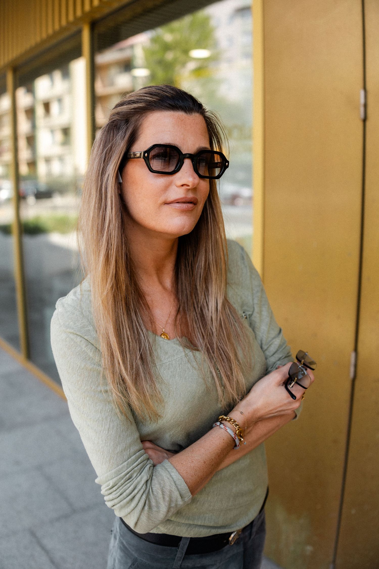 Person in gray sweater and sunglasses posing against gold wall in outdoor photoshoot.