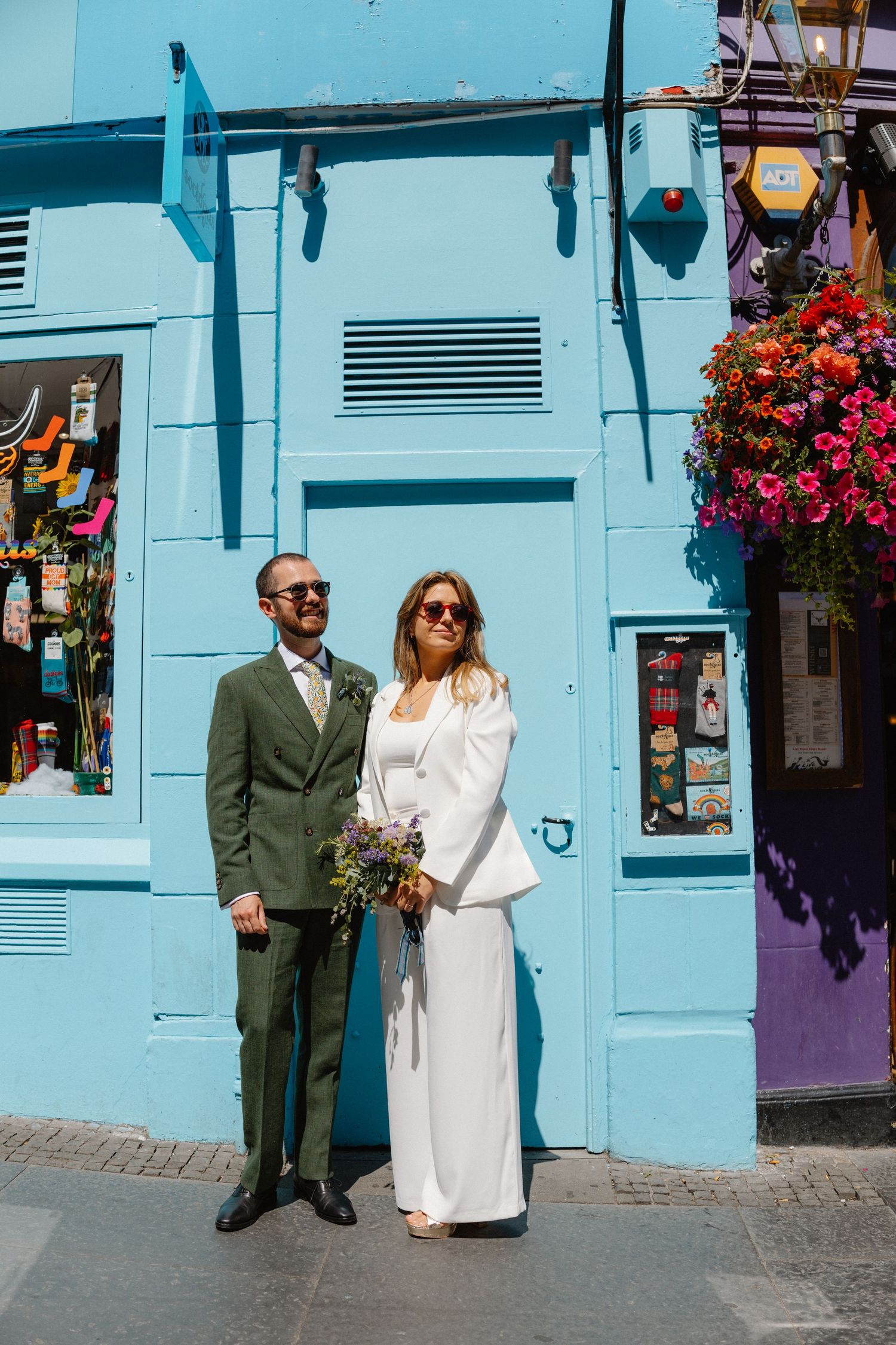 Couple standing against vibrant turquoise blue wall with hanging flower baskets.