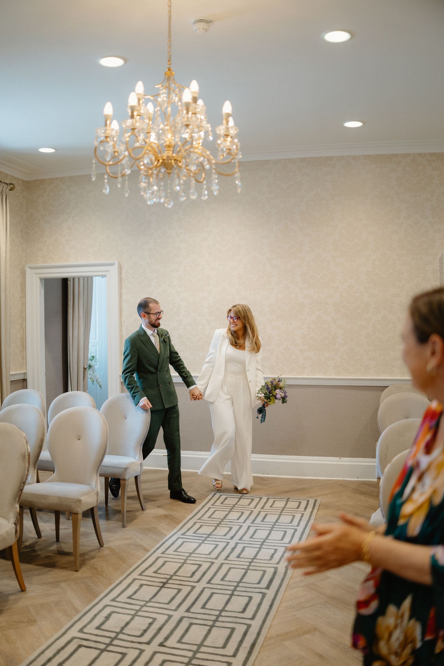 Newlywed couple walks hand in hand down aisle under gold chandelier in elegant ceremony room.