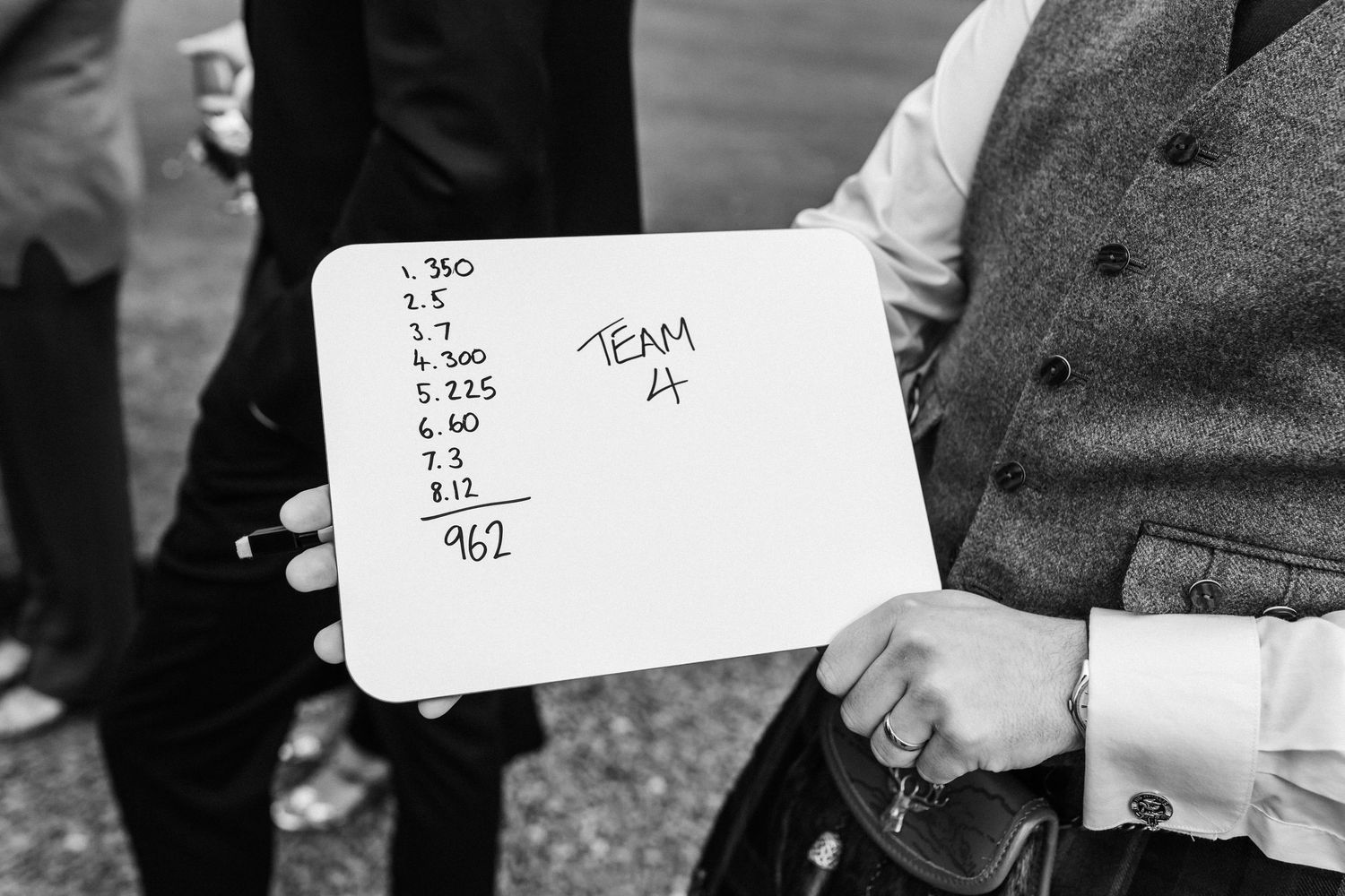 A whiteboard displaying team scores during outdoor wedding lawn games with a guest holding it.