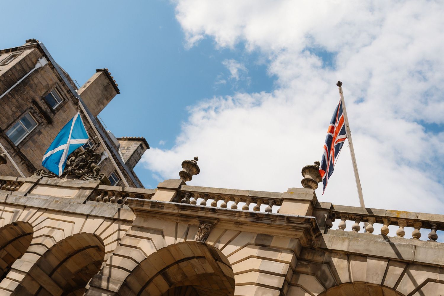 A Scottish Saltire and Union Jack flag flying above a historic stone building with ornate balustrade against a blue sky.