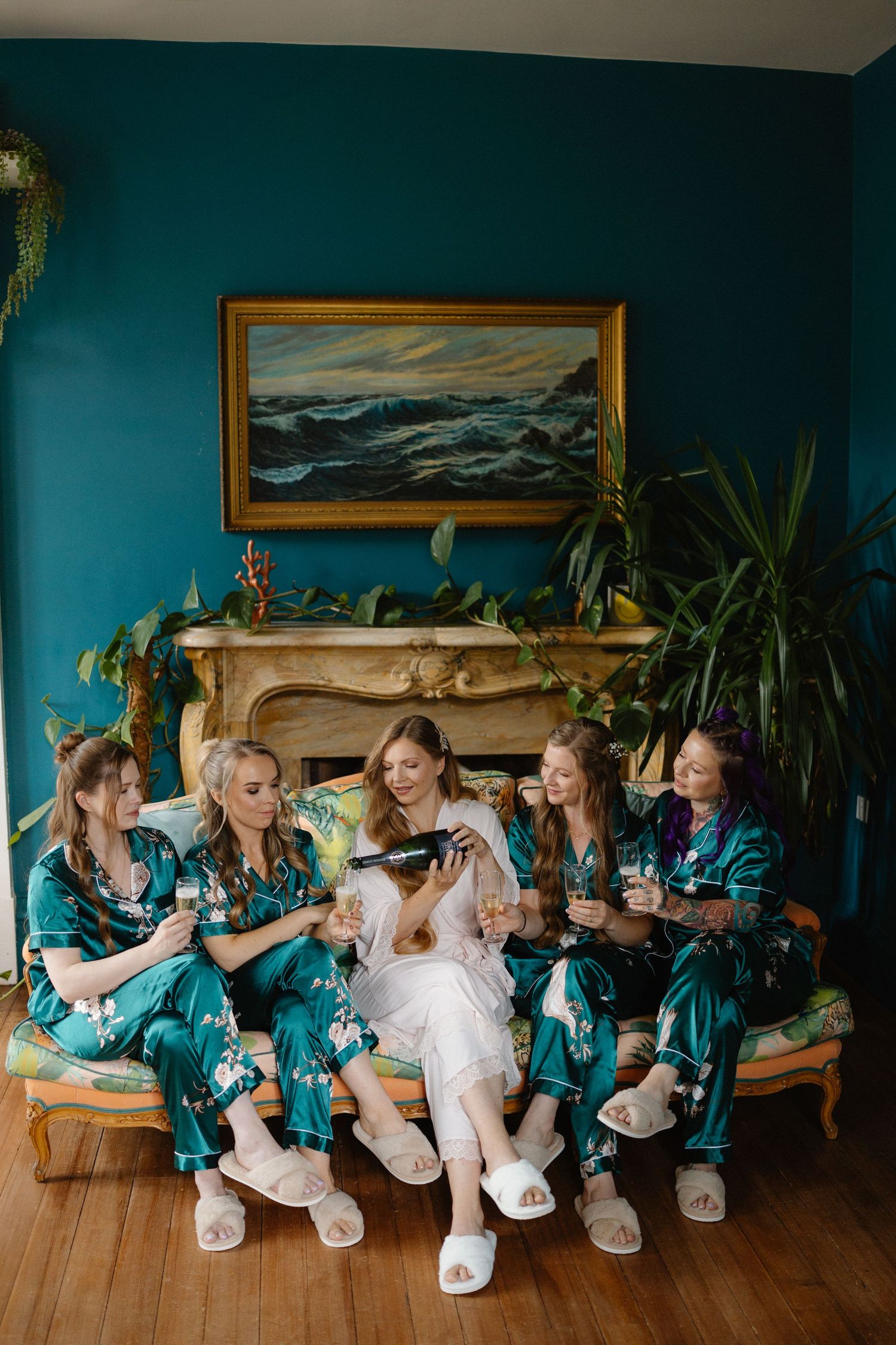 Bridesmaids in teal satin robes toast champagne on a couch before the wedding ceremony.