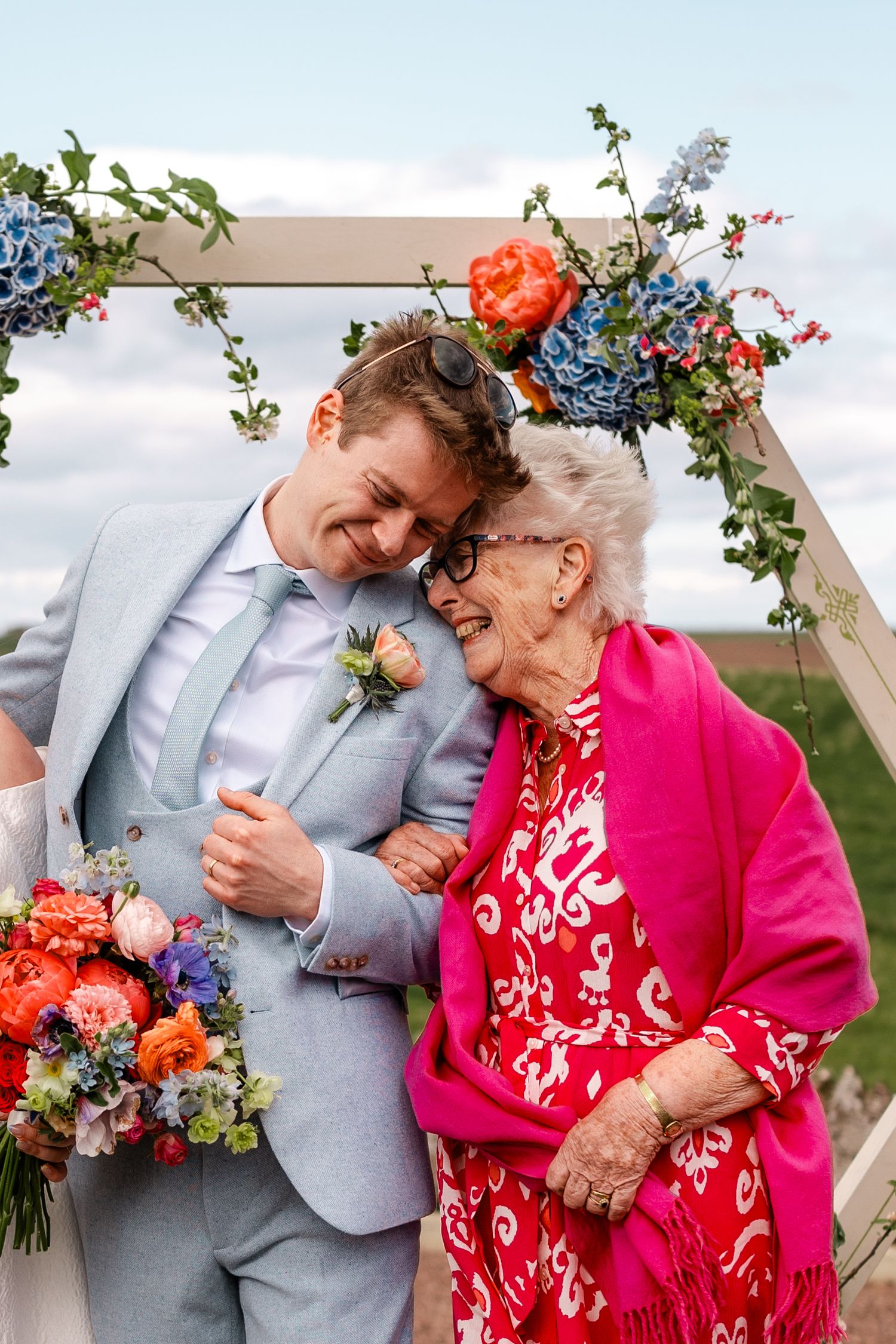 Groom in light blue suit shares a joyful moment with elderly woman in pink shawl at floral wedding arch.