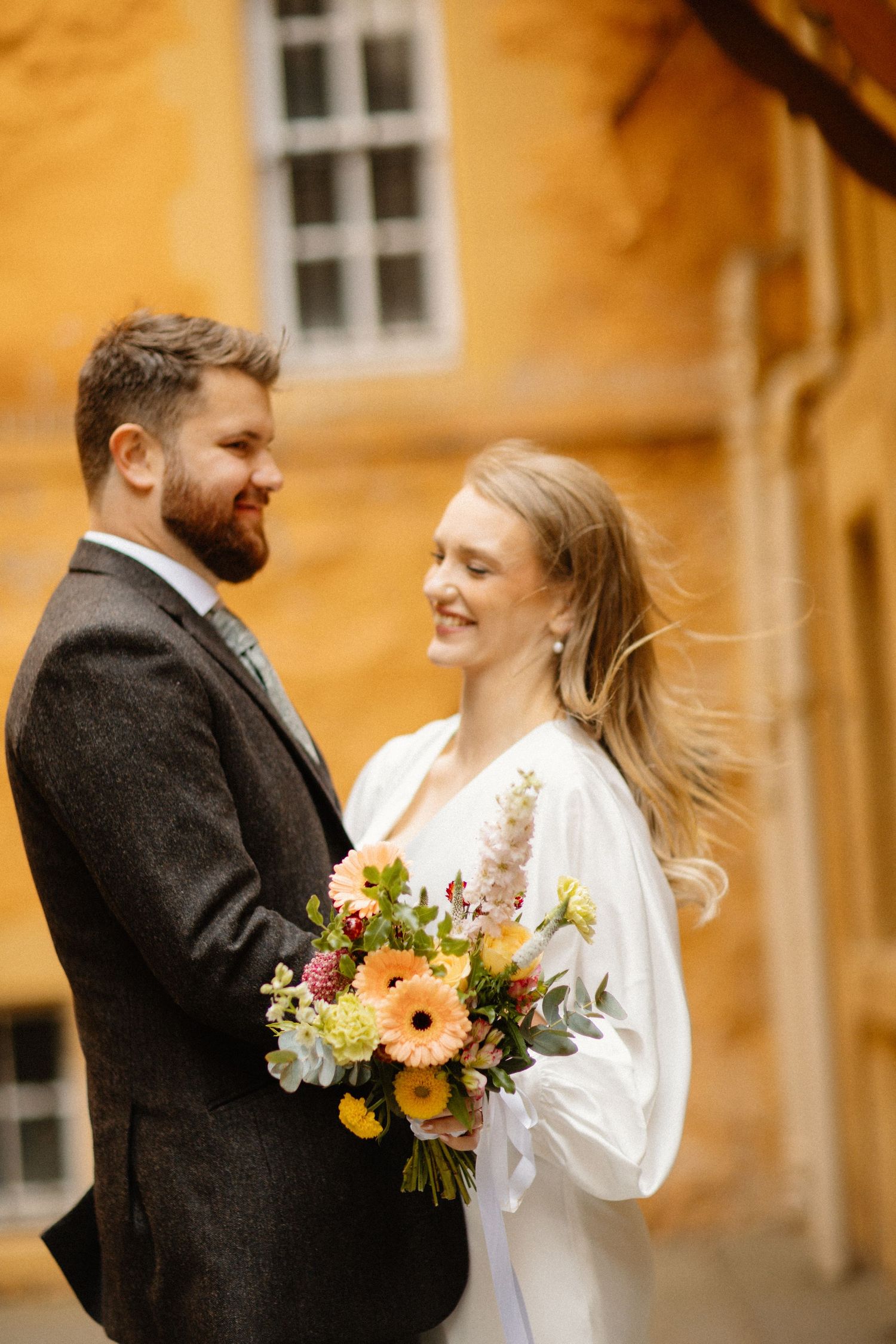 Smiling bride holds colorful bouquet of gerberas beside bearded groom in grey suit against yellow wall.