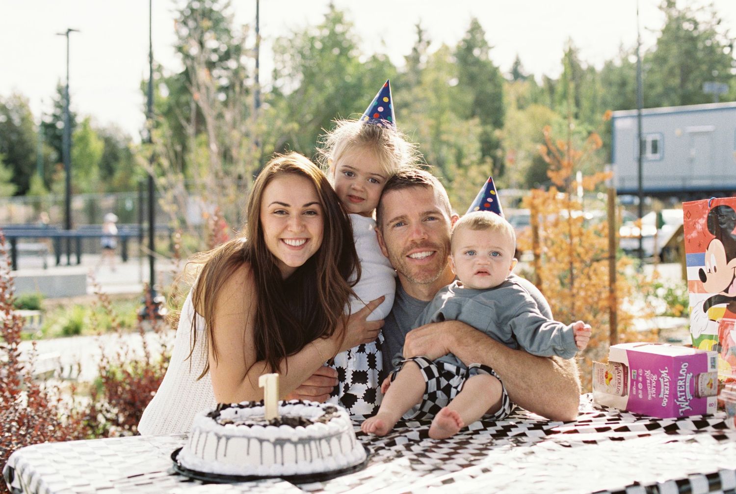 Family celebrating birthday outdoors with cake and party hats, parents smiling with two young children at decorated table.