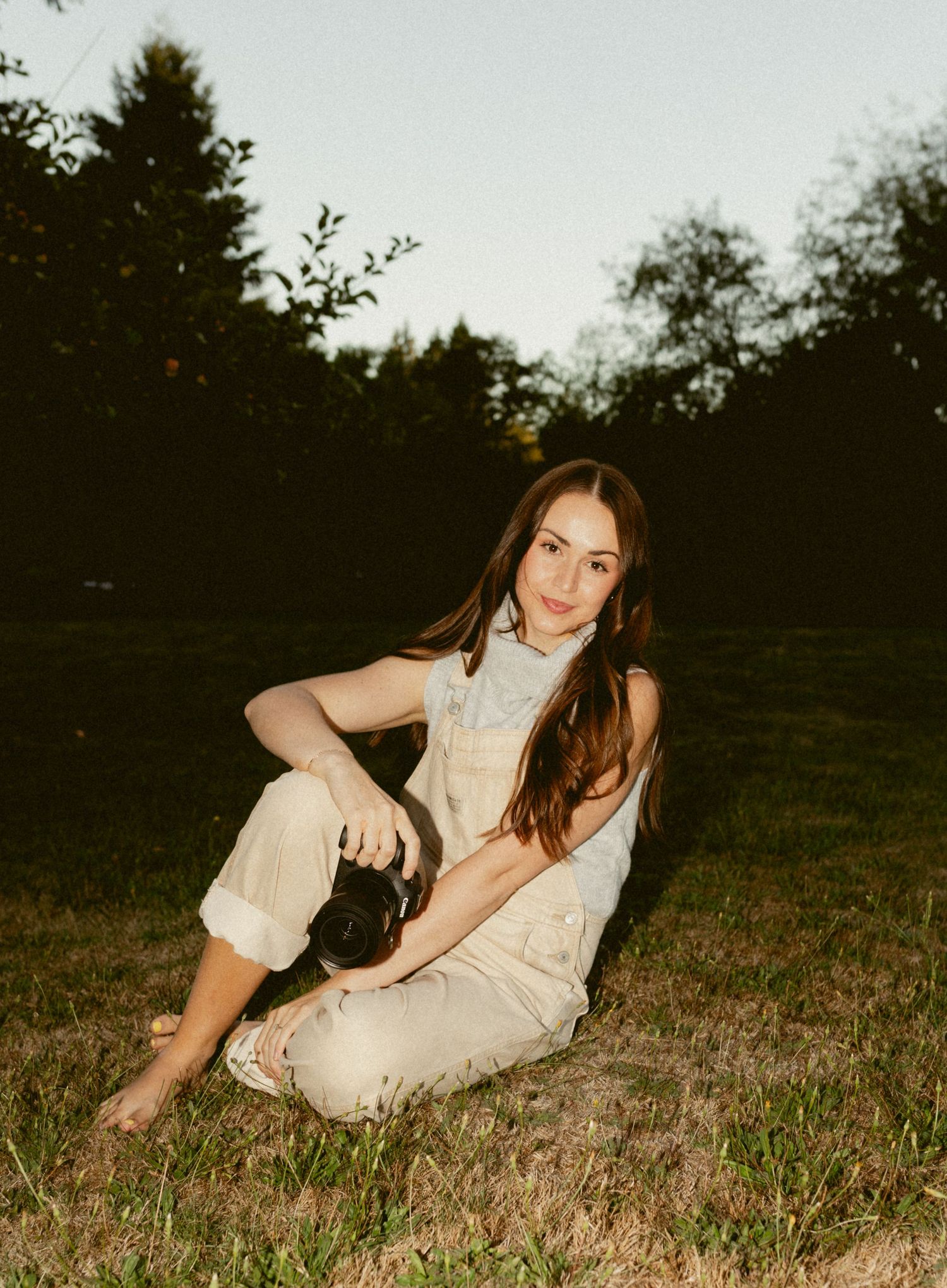 Someone in a cream-colored outfit sits on grass at dusk with trees silhouetted in the background.