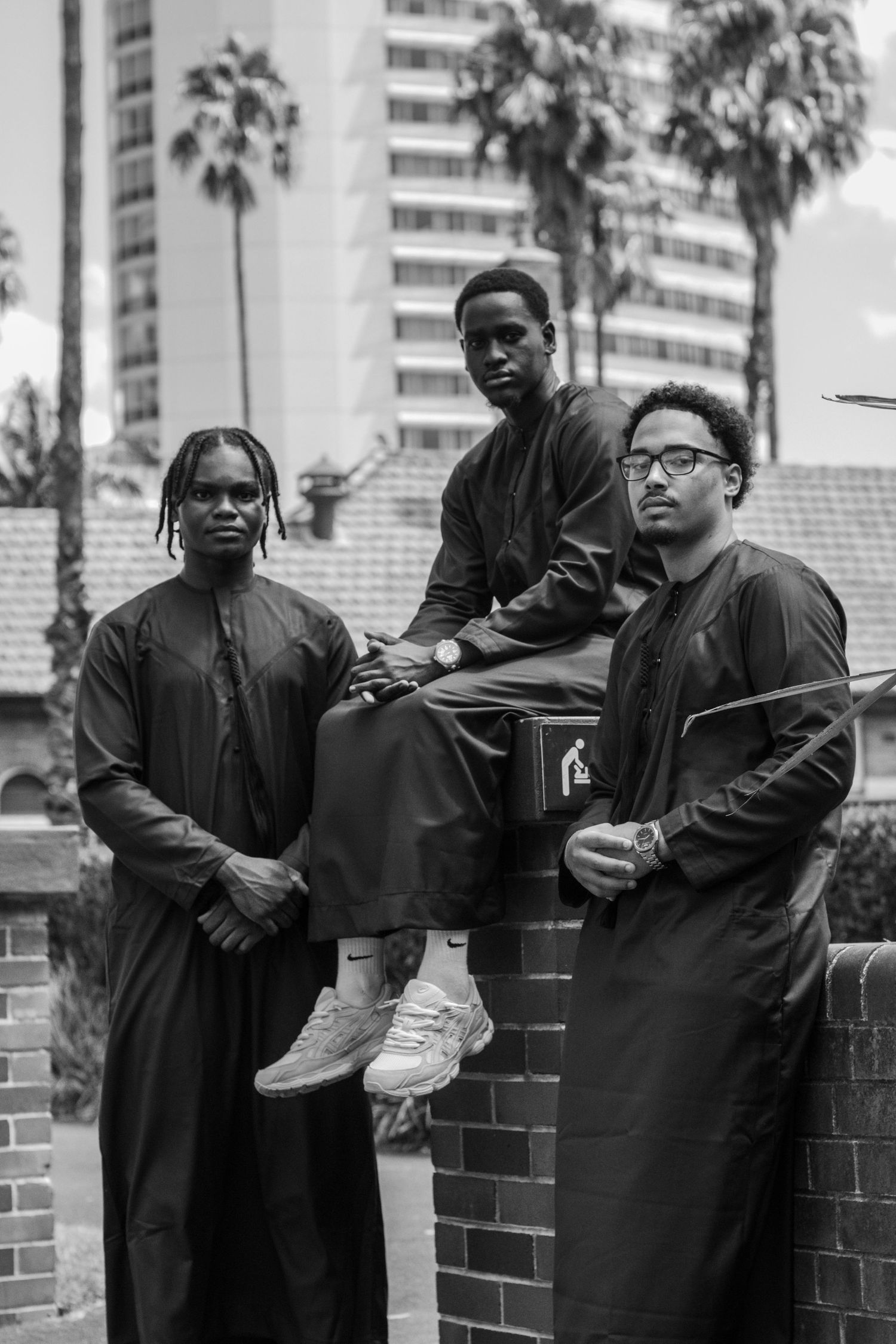 Black and white portrait of three men in traditional thobes posing near brick wall with palm trees.