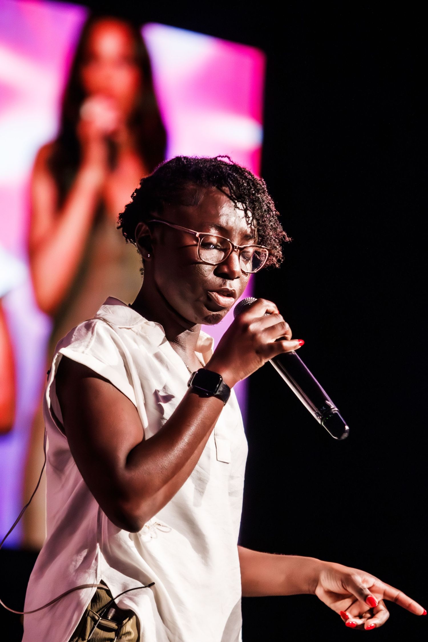 Young woman with glasses performs spoken word into microphone on stage.