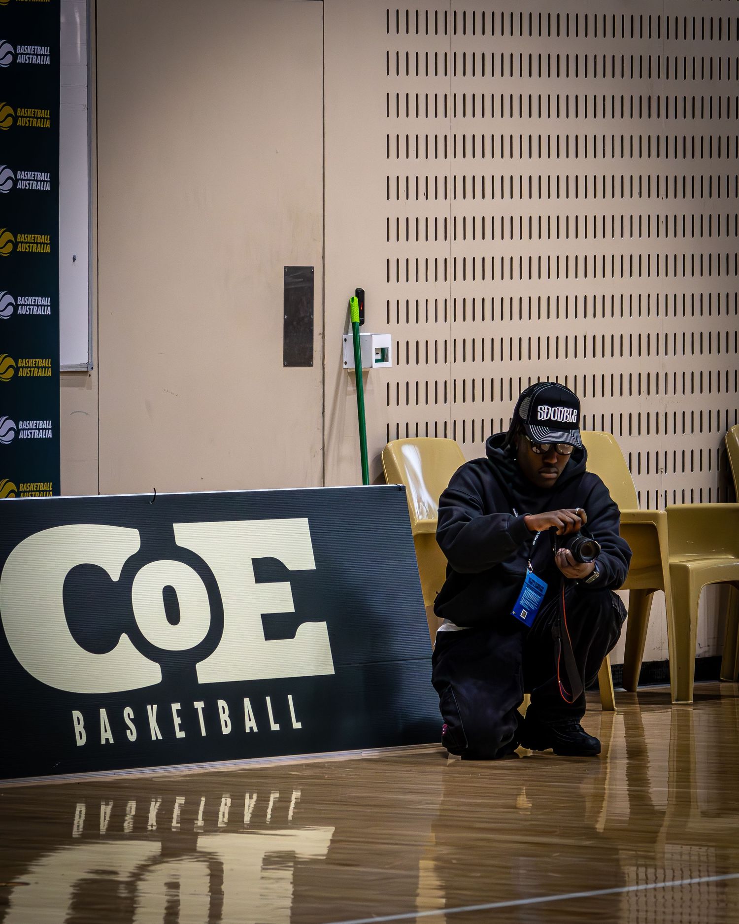 Basketball coach crouches beside CoE Basketball signage checking phone at indoor court.
