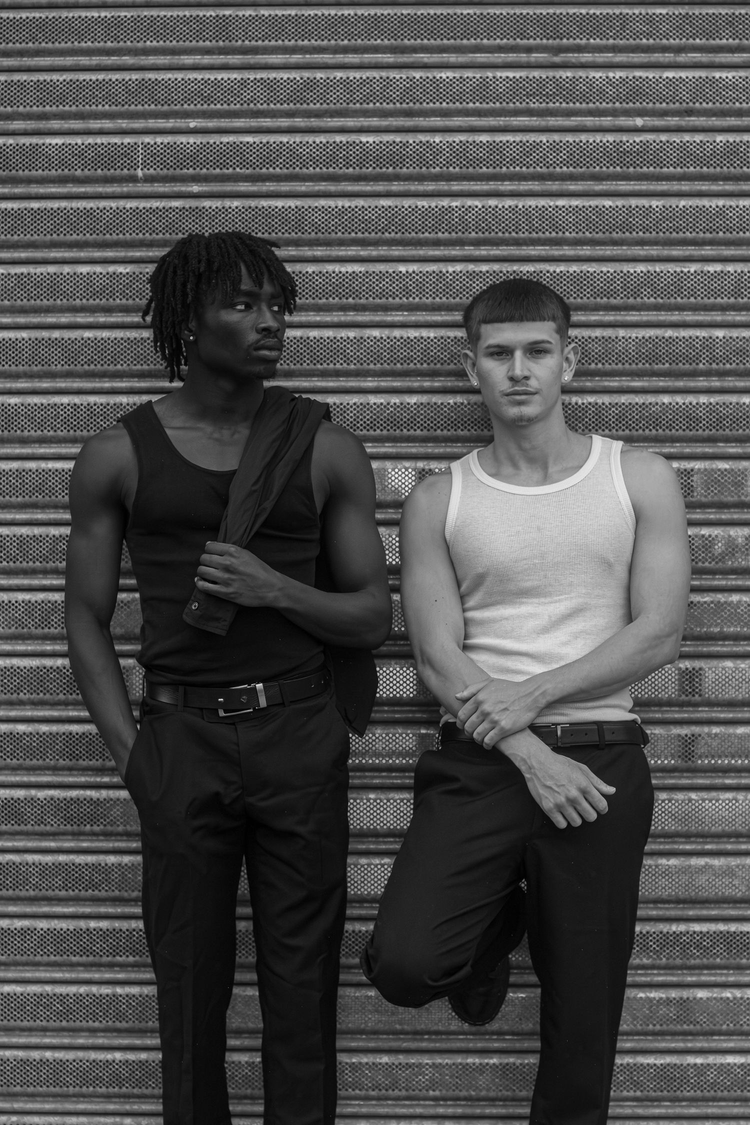 Black and white portrait of two men in tank tops leaning against corrugated metal shutter wall.