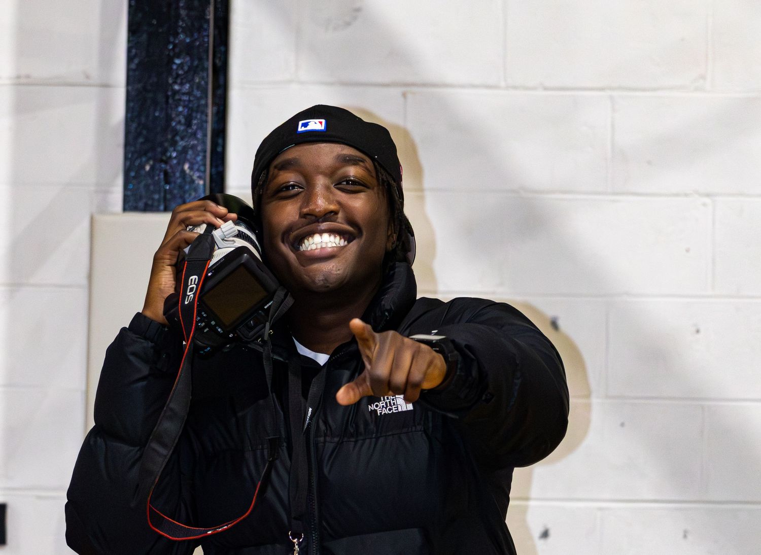 Smiling young man holds camera and points at viewer wearing black puffer jacket and cap.