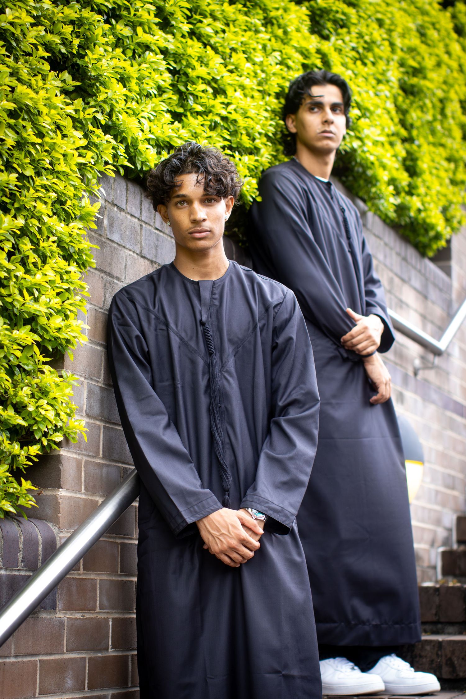 Two young men in dark traditional thobes pose on outdoor stairs with green foliage wall backdrop.