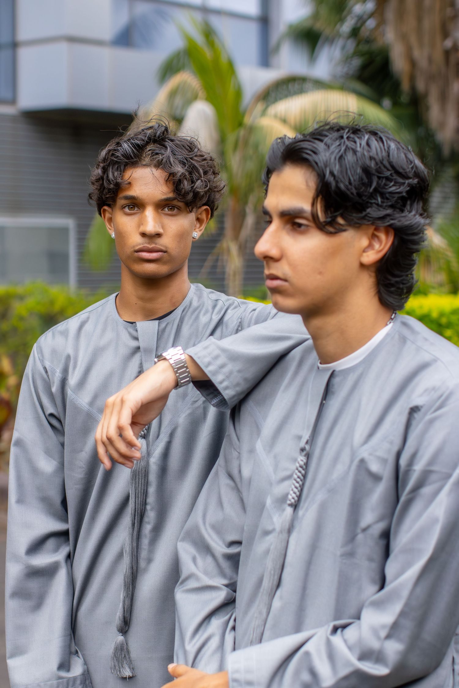 Two young men in matching grey traditional thobes pose outdoors with palm trees behind them.