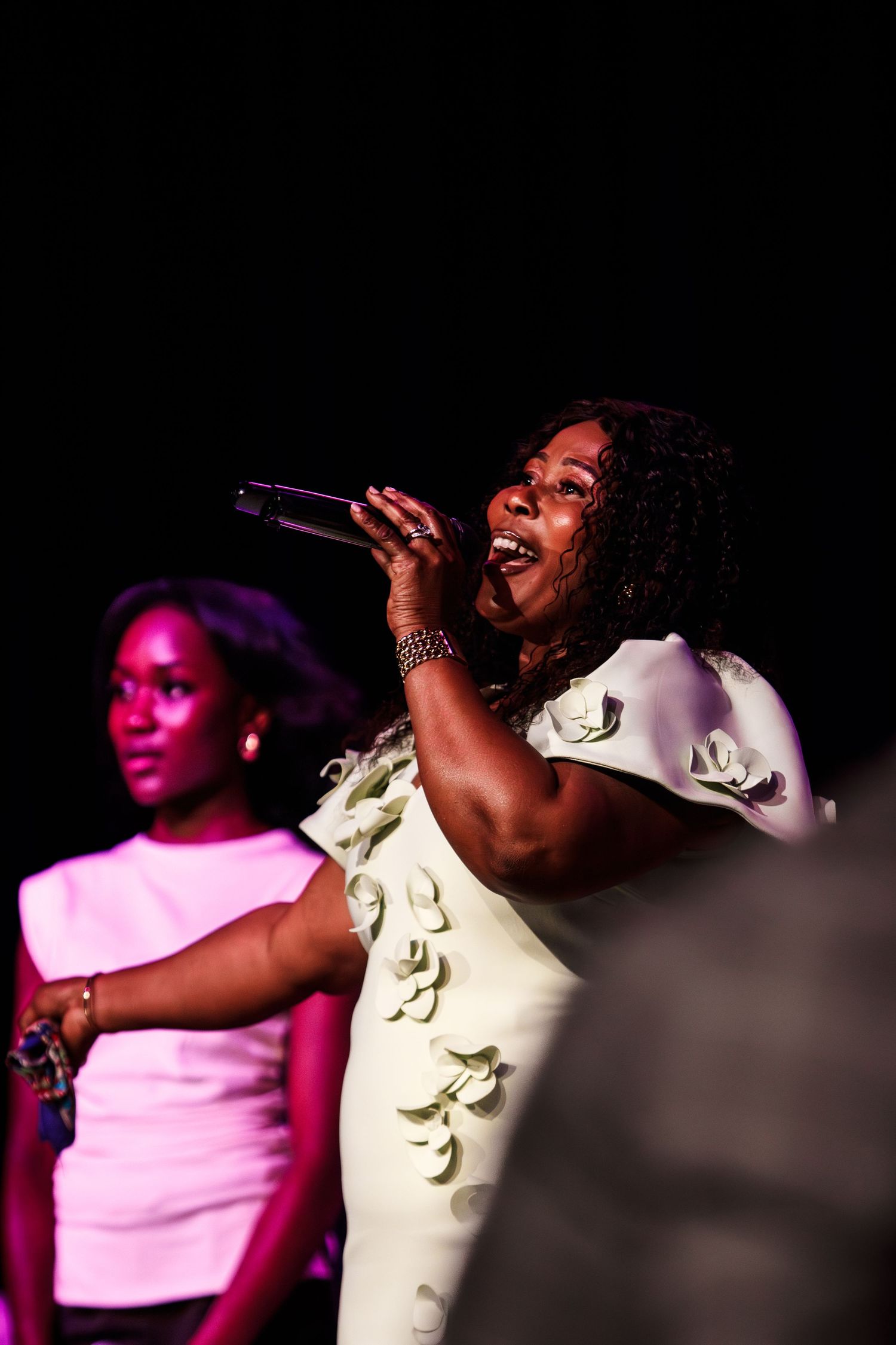Woman in white floral dress performs on stage holding microphone with singer in background.
