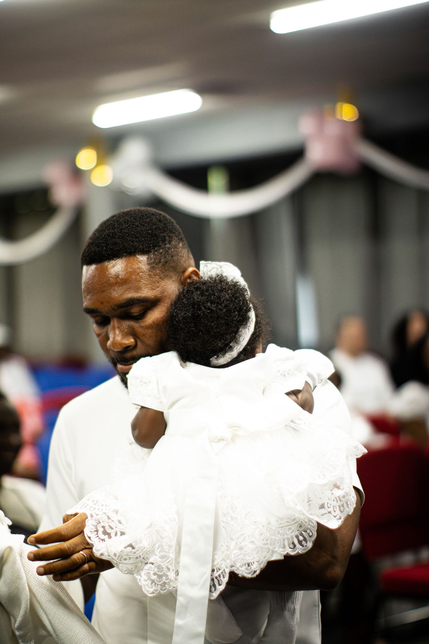 Man in white shirt tenderly holds a baby dressed in white lace christening gown at ceremony.