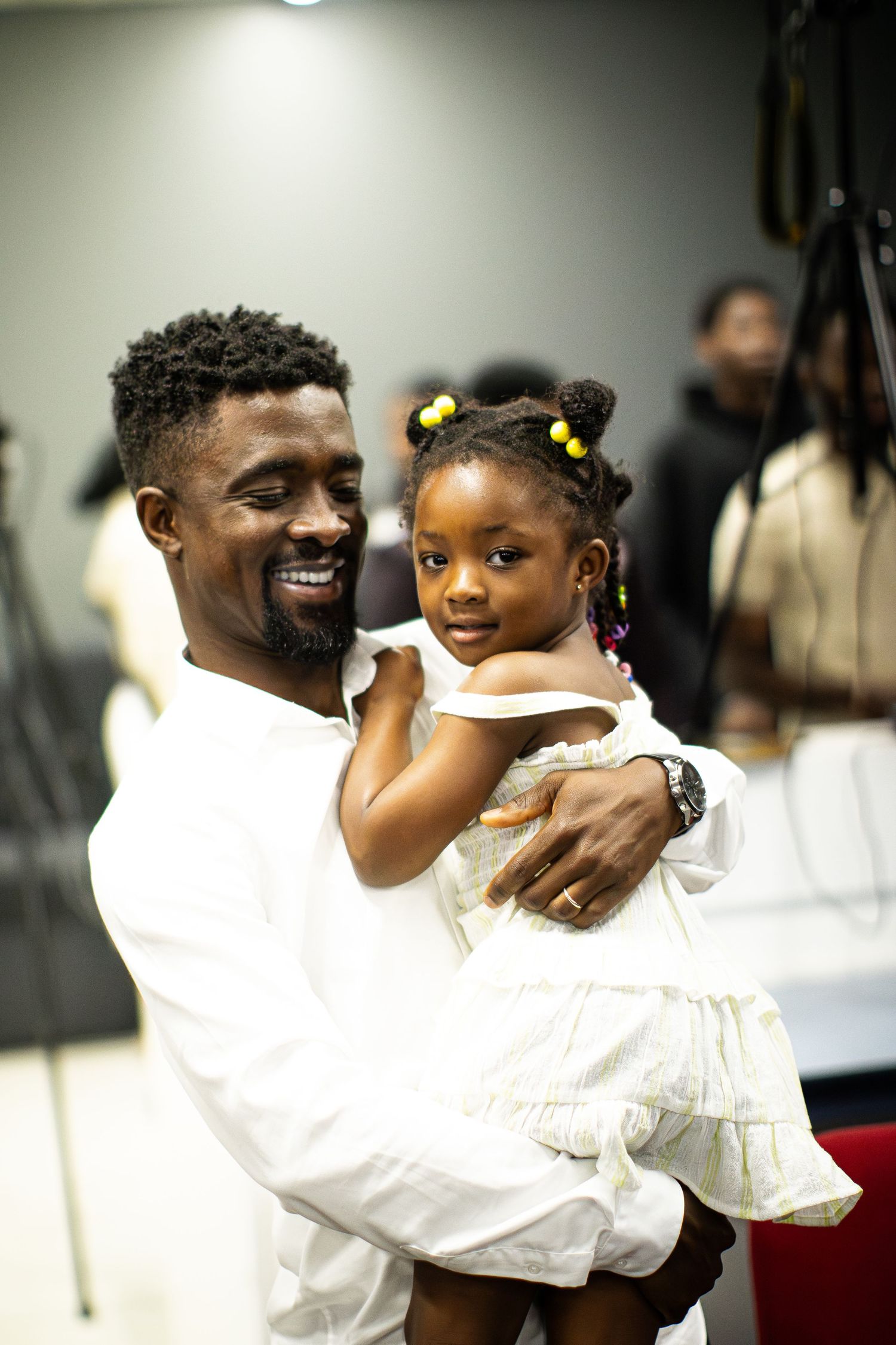 Smiling father holds young daughter with colorful hair accessories at indoor event.