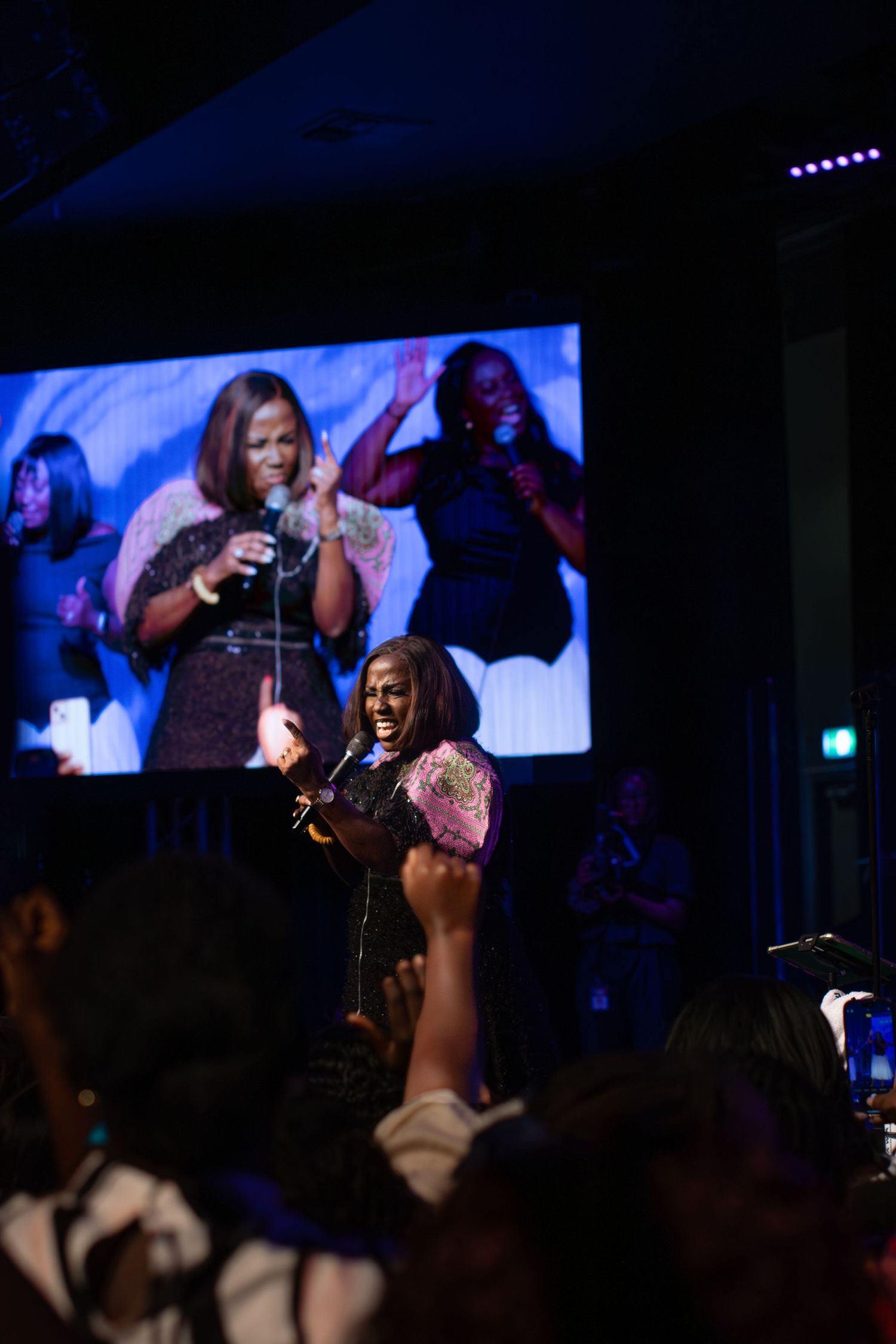 Female gospel singer performs on stage with live screen projection behind her.