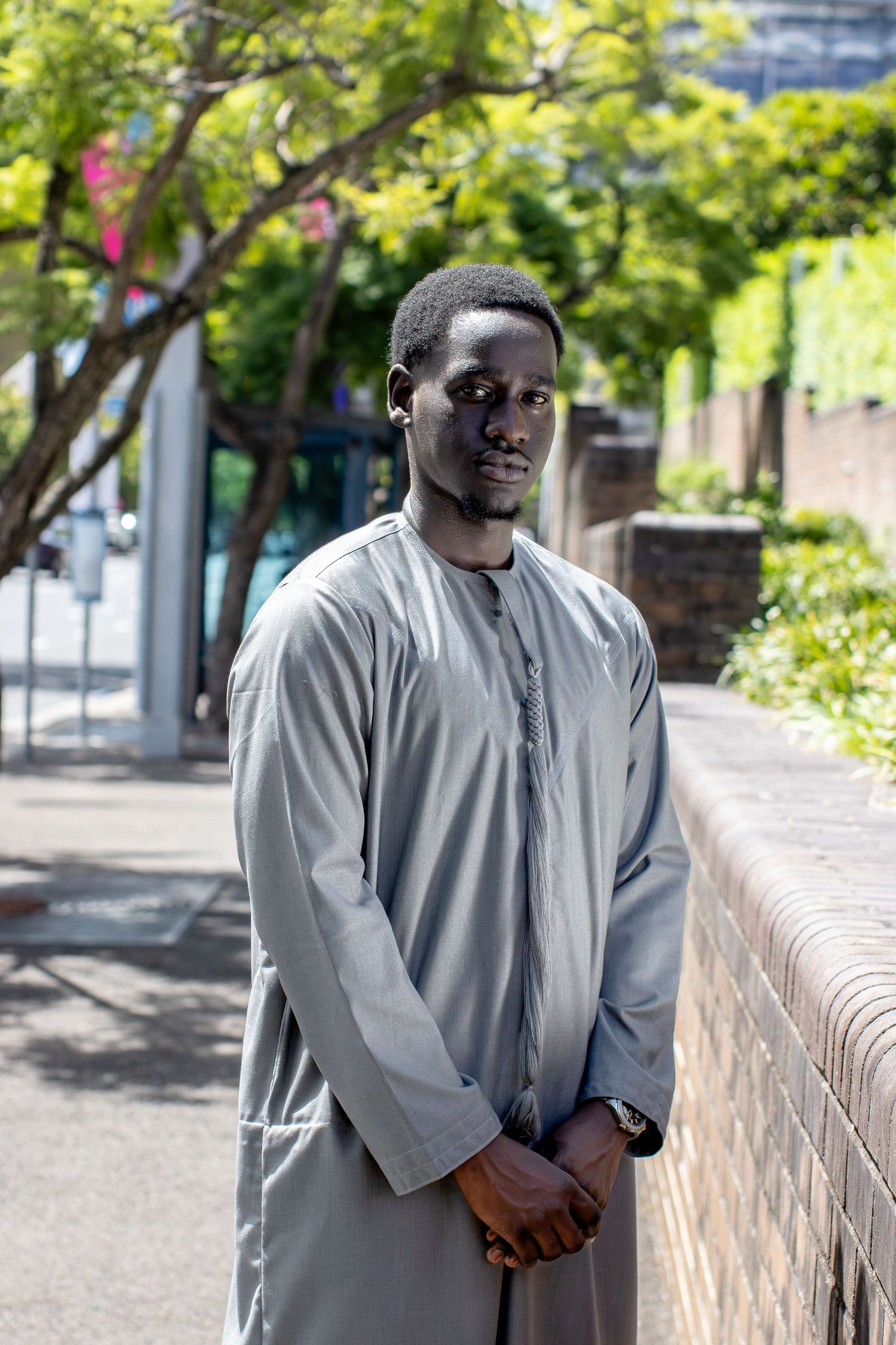 Man in grey traditional thobe stands on sunny urban sidewalk with trees and wall behind him.