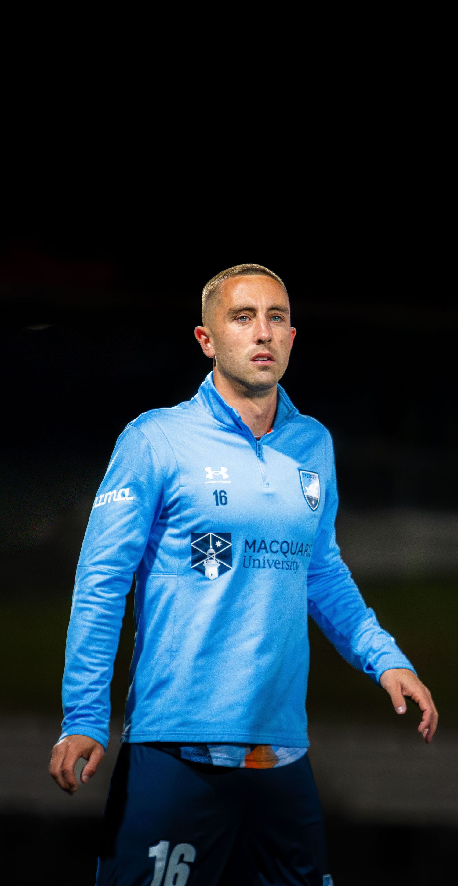 Male soccer player #16 in blue Macquarie University sponsored Under Armour jersey during a night match.