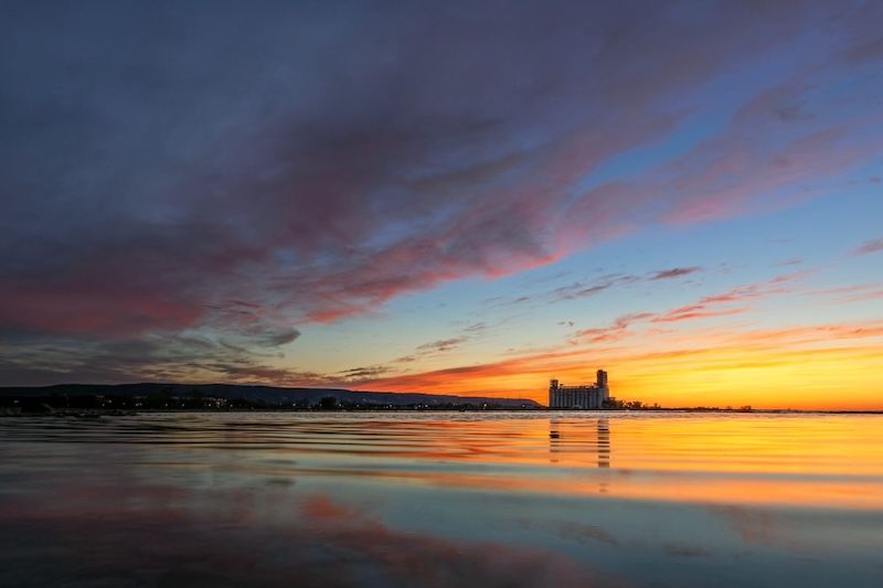 Vivid pink and orange sunset reflecting on calm water with a grain elevator silhouette on the horizon.