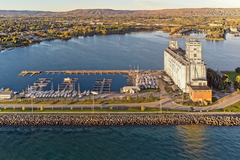 Aerial view of a marina and grain elevator on a calm waterfront with a rocky breakwall and colorful autumn trees.