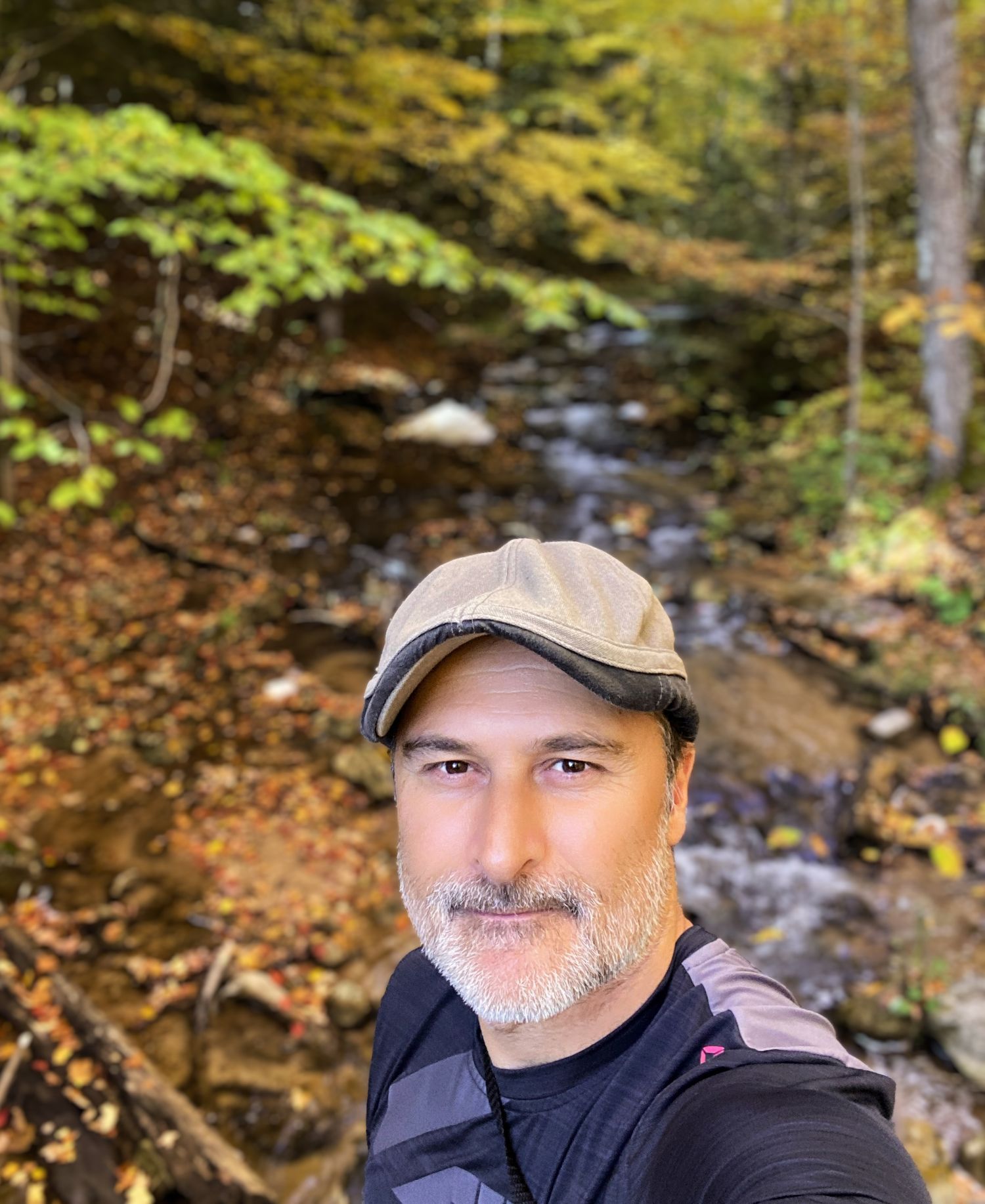 Man in cap takes selfie beside rocky autumn forest stream with colorful foliage background.