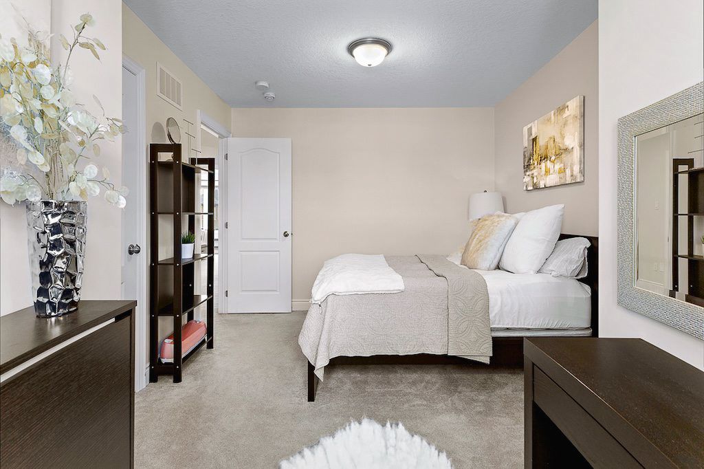 Cozy carpeted bedroom with neutral tones, dark wood furniture, decorative bookshelf, and soft lighting.