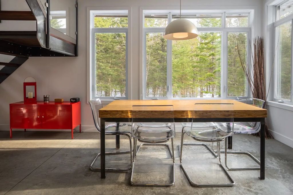 Contemporary dining area with rustic wood table, ghost chairs, large windows overlooking snowy forest.