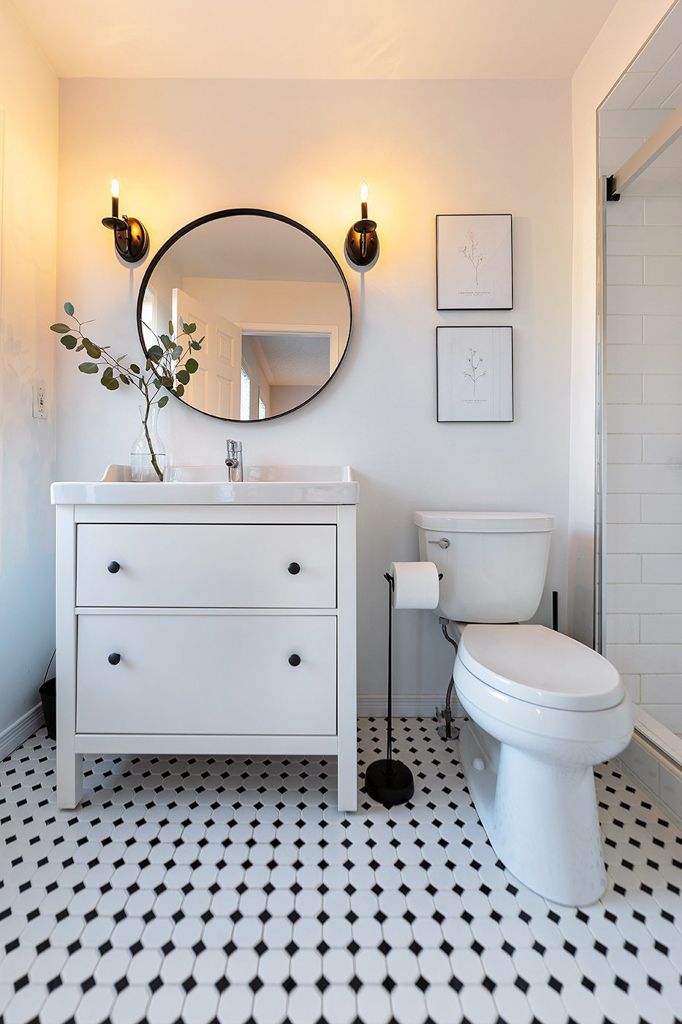 Elegant white bathroom with round mirror, black sconce lights, white vanity, and classic black and white tile floor.