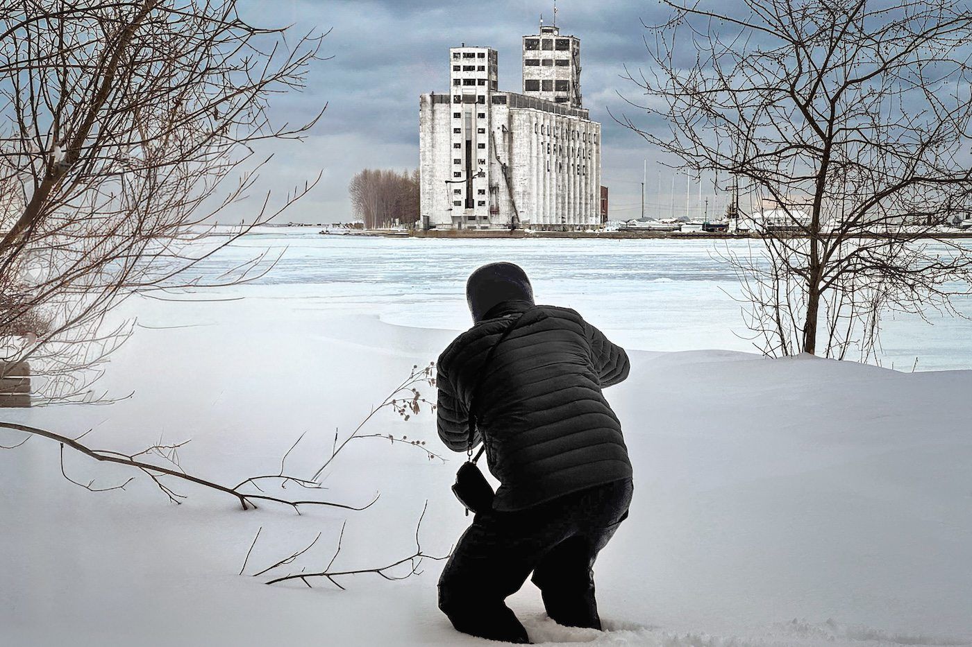 Photographer kneeling in snow capturing an industrial grain elevator building in winter.