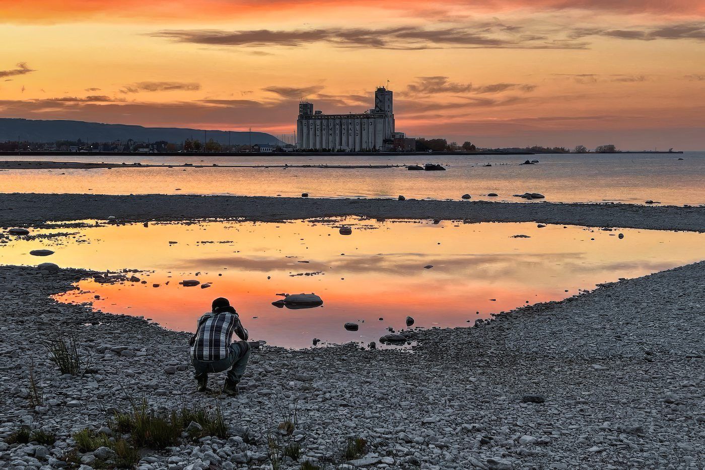 Photographer crouching on rocky shore at sunset with a grand building reflected in water.