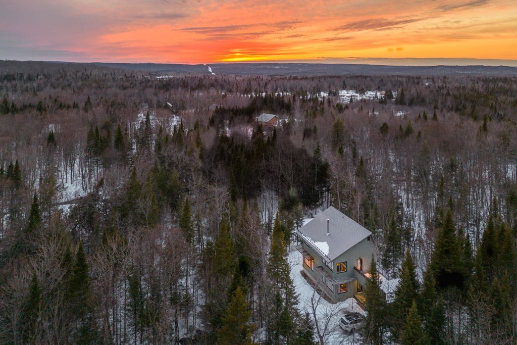 Aerial winter sunset view of an isolated house glowing warmly amid dense snow-covered forest.