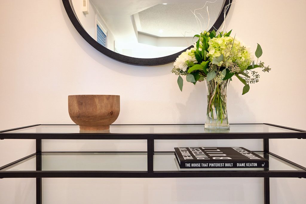 Black metal console table with round mirror, wood bowl, flowers, and coffee table books.