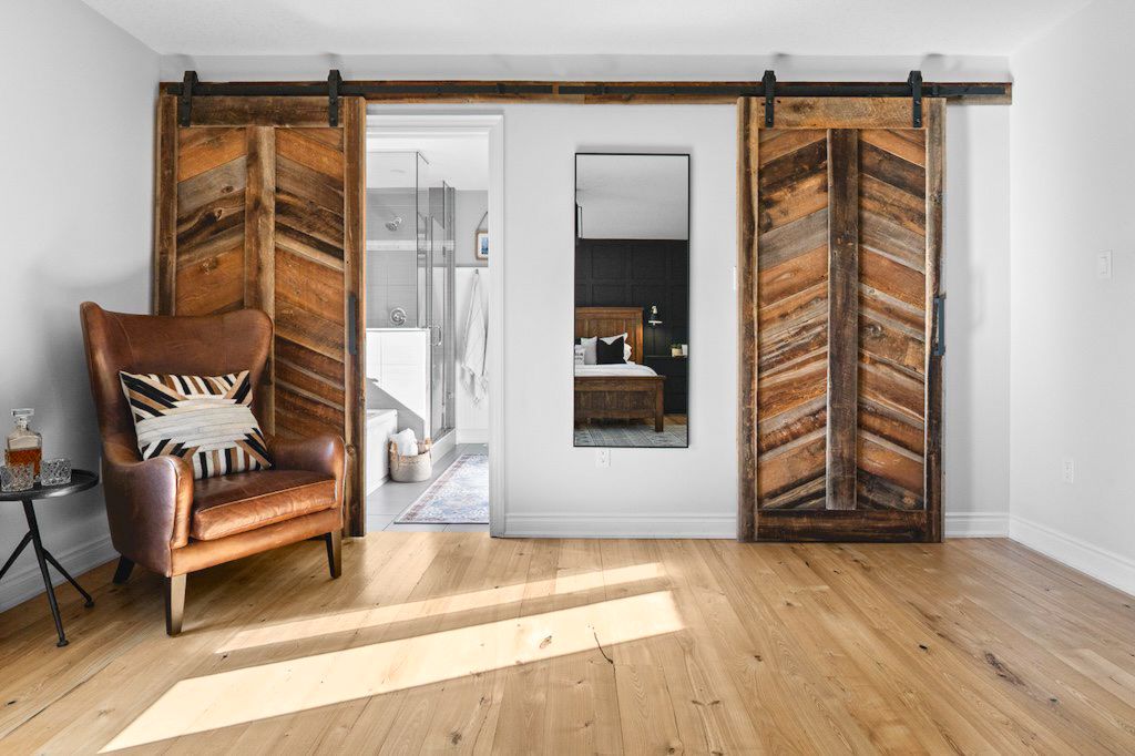 Rustic reclaimed wood sliding barn doors open to reveal a bedroom, with a leather armchair on light hardwood floors.